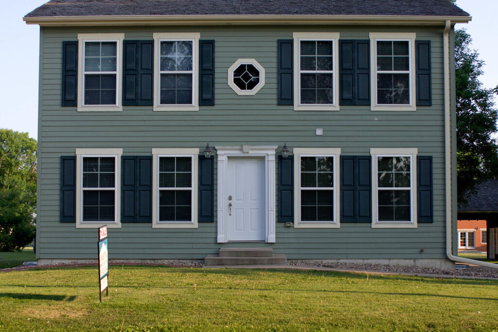 A grey two-storey colonial home with dark shutters and a sign on the front lawn